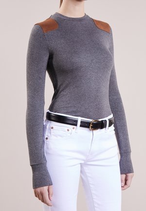 Femme portant un haut gris ajusté à manches longues avec des renforts d’épaule marron, un pantalon blanc et une ceinture noire avec une boucle dorée.