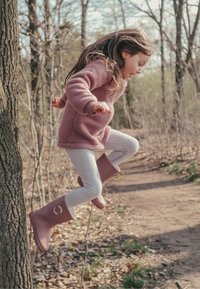 Bottes en caoutchouc roses avec un accent incurvé, leggings gris et un pull polaire rose clair, alors qu'un enfant saute d'un arbre dans une zone boisée.