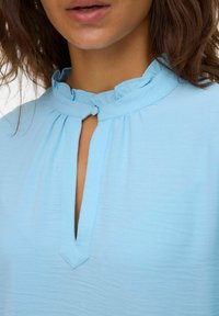 Donna che indossa una blusa blu chiaro con colletto arricciato e una chiusura a un solo bottone sopra un scollo a V.