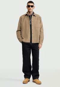 Uomo con occhiali da sole neri, giacca a bottoni color crema, pantaloni neri, sciarpa a fantasia e stivali color crema, in piedi con le mani in tasca.