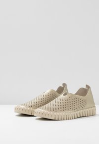Zapatos de tela beige sin cordones con parte superior perforada, suela flexible y bordes texturizados estriados. Diseño minimalista sin herrajes o acentos visibles.