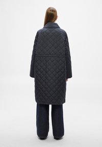 Πανωφόρι από μαύρο καπιτονέ ύφασμα με γιακά, μακριά μανίκια και άνετη γραμμή, που διαθέτει μοτίβο ρόμβου και λεπτομέρεια με ραφή στην πλάτη.