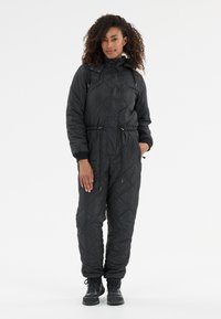 Zwart gewatteerd jumpsuit met hoge kraag, koordjes in de taille en ritszakken. Heeft elastische manchetten en wordt gecombineerd met zwarte laarzen.
