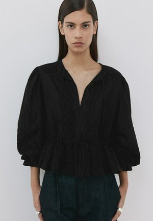 Joven con cabello largo y oscuro que lleva una blusa negra bordada con mangas abullonadas y pantalones oscuros de cintura alta, de pie frente a un fondo blanco.