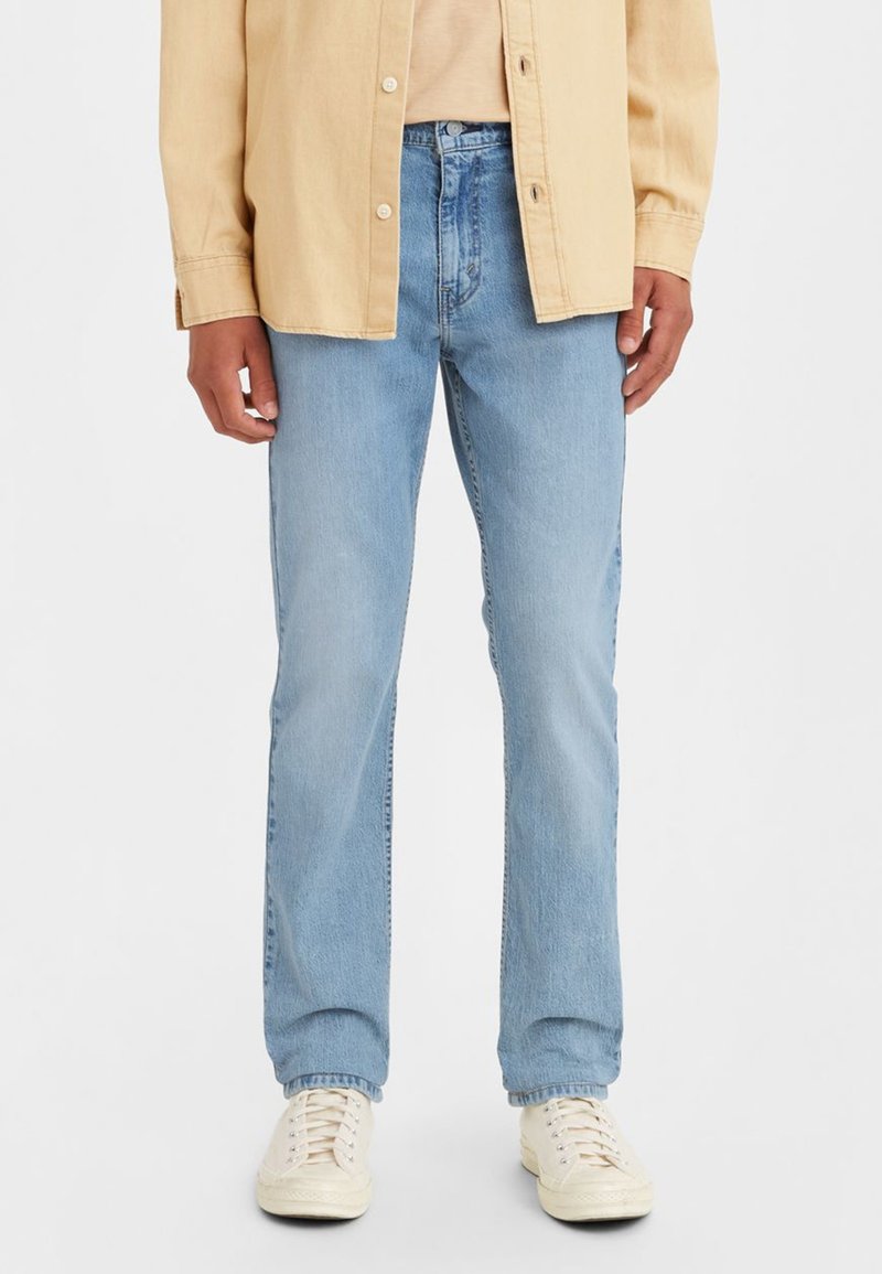 Jeans de mezclilla azul claro con corte recto, que presentan un diseño clásico de cinco bolsillos y una sutil textura deslavada. Combinados con una camisa beige.