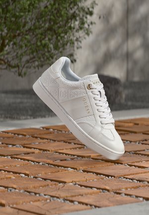 Baskets blanches Guess avec motif en relief et logo doré, flottant au-dessus d'un sol extérieur en bois et pierre avec un arrière-plan de verdure floue.