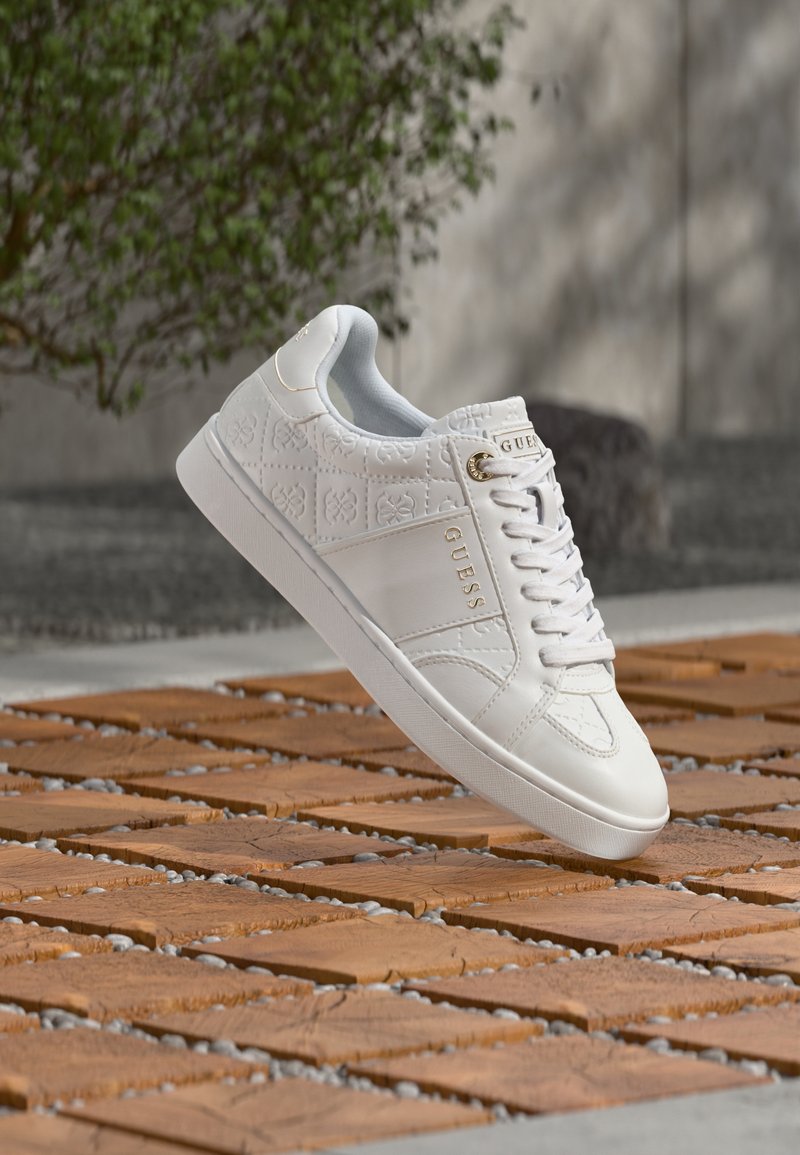 Biela teniska Guess s reliéfnym vzorom a zlatým logom, vznášajúca sa nad drevenou a kamennou vonkajšou podlahou s rozmazaným zeleným pozadím.