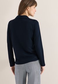 Femme aux cheveux châtain mi-longs portant un haut bleu marine à manches longues et un pantalon gris, tournée vers un mur beige, vue de dos et de profil.