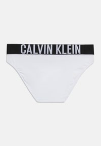 Λευκό εσώρουχο στυλ μπικίνι με μαύρη ζώνη μέσης που φέρει μεγάλα λευκά γράμματα "CALVIN KLEIN" στο εμπρόσθιο μέρος.