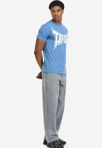 Blaues Baumwoll-T-Shirt mit großem weißem "TAPOUT"-Logo, kombiniert mit grauer Jeanshose und schwarzen Sportschuhen. Das Modell steht seitlich.