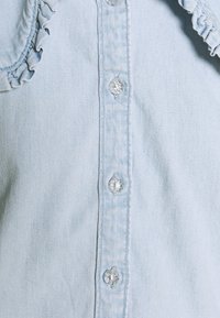 Chemise en denim bleu clair avec bords à volants, boutons transparents et texture unie, mettant en valeur le design décontracté classique.