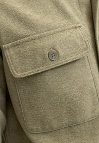 Olivengrønt ullstoff med teksturert overflate, med en lomme som har en stor, rund, firehulls knapp for lukking.