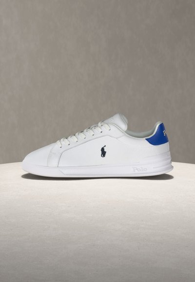 Λευκό χαμηλό αθλητικό παπούτσι με μπλε ταμπελάκι στη φτέρνα, μαύρο λογότυπο Polo στο πλάι, λευκά κορδόνια και λαστιχένια σόλα που ακουμπά σε φωτεινή επιφάνεια.