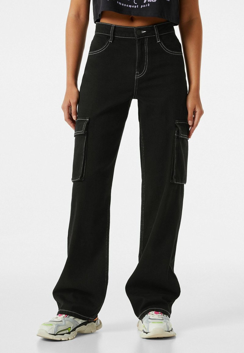 Svarta cargobyxor med kontrasterande vita sömmar, utrustade med två sidfickor och en avslappnad passform; bärs med grå och gröna sneakers.