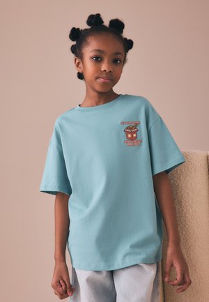Ung pige med stylet hår iført en lyseblå T-shirt med en ramen-grafik og tekst, stående mod en ensfarvet baggrund.