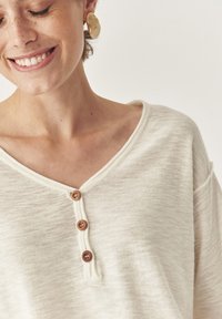 Helles beiges V-Ausschnitt-Shirt mit strukturiertem Stoff, drei Holzknöpfen am Ausschnitt und lockerem Schnitt mit abgesenkten Schultern.