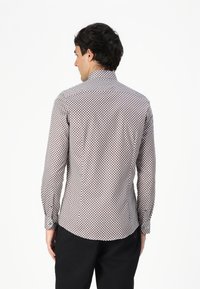 Chemise à manches longues blanche avec un motif géométrique rouge et marine, dotée d'un col haut et de poignets boutonnés, fabriquée dans un tissu léger.