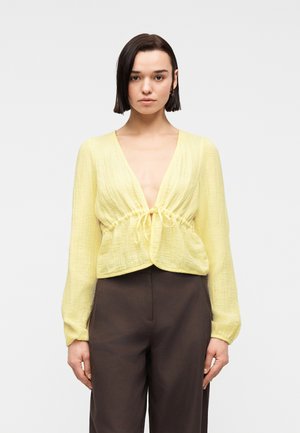 Femme aux cheveux courts et foncés portant un chemisier jaune texturé à manches longues noué à la taille et un pantalon marron foncé taille haute.