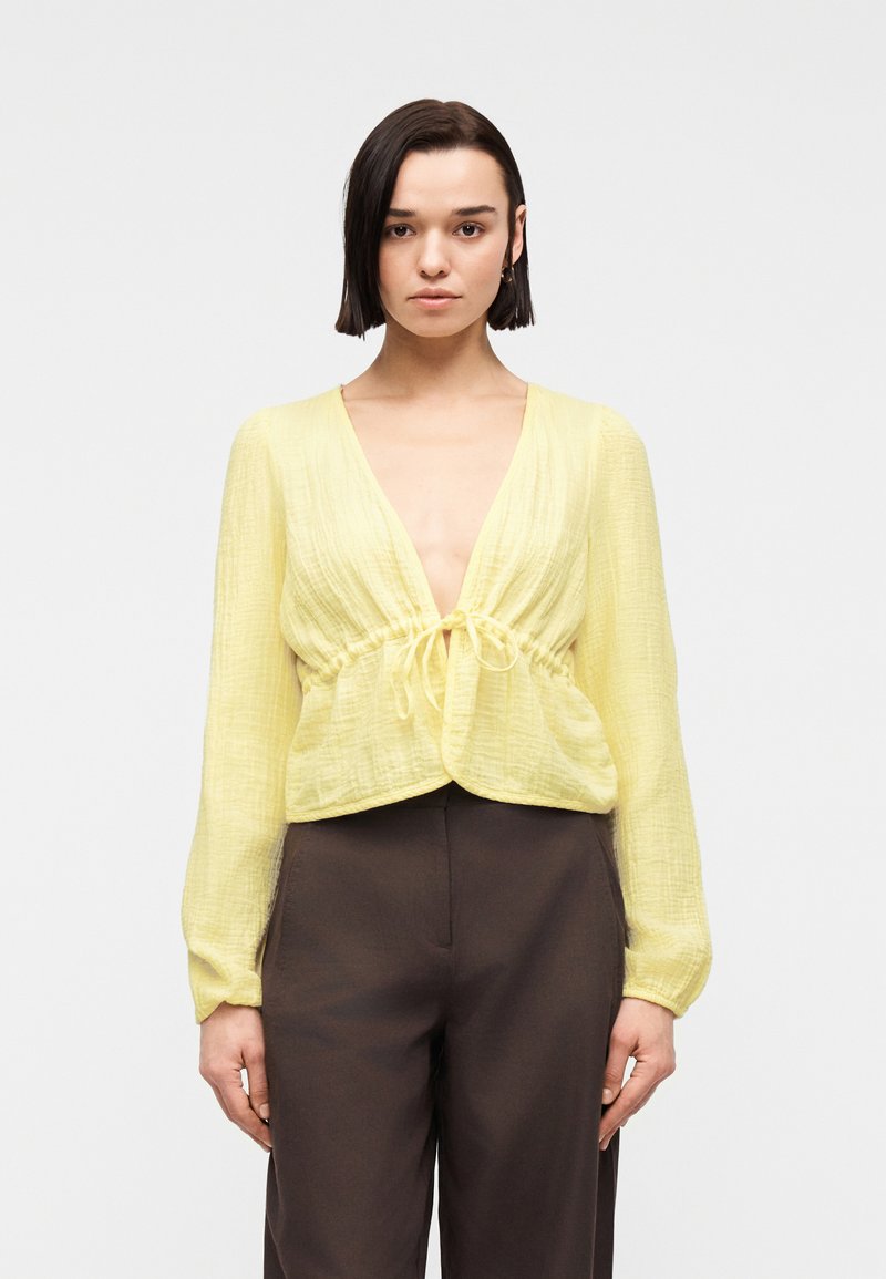 Femme aux cheveux courts et foncés portant un chemisier jaune texturé à manches longues noué à la taille et un pantalon marron foncé taille haute.