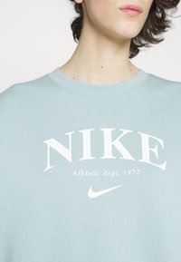 Detailní záběr na osobu v světle modré mikině Nike s bílým nápisem "NIKE Athletic dept. 1972" a logem Nike swoosh.
