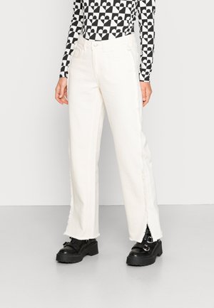 Jeans brancos de perna larga com bainhas desfiadas, apresentando uma cintura alta e fendas laterais, combinados com sapatos pretos robustos e uma blusa de manga longa estampada.