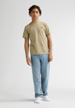 T-shirt in cotone color kaki con scollo rotondo, maniche corte e dettaglio stampato sul petto, abbinata a jeans a gamba dritta azzurri chiari e sneakers bianche.