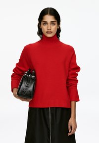 Frau trägt einen roten Rollkragenpullover und einen schwarzen Rock und hält eine schwarze Lederhandtasche vor einem weißen Hintergrund.