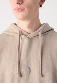Béžová mikina s kapucňou vyrobená z ribbed látky, ktorá má stiahnuťeľnú kapucňu a textúrované kruhové logo na hrudníku.