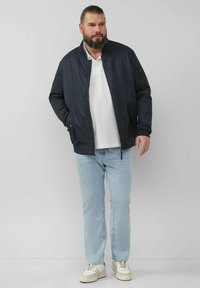 Marineblauer Bomberjacke mit Reißverschlussfront, gerippten Bündchen und seitlichen Taschen; kombiniert mit hellblauen Jeans und cremefarbenen Sneakers.