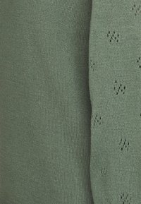 Pull vert tricoté avec une texture douce, présentant des motifs décoratifs ajourés éparpillés sur le tissu. Encolure simple et arrondie.