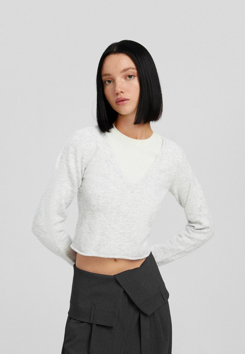 Helles graues, kurzes Sweater mit langenÄrmeln und tiefem V-Ausschnitt, über einem weißen Oberteil getragen. Der Stoff hat eine weiche Textur. Dunkelgrauer, hochtaillierter Rock.