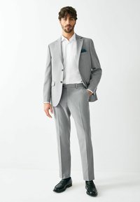 Traje gris a medida con chaqueta ajustada y pantalones a juego. Camisa blanca impecable debajo, acentuada con un pañuelo de bolsillo oscuro. Zapatos de vestir negros.