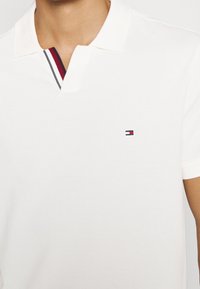 Polo bianca con colletto a due bottoni, caratterizzata da dettagli a righe blu navy e rosse. Include un piccolo logo ricamato sul petto.