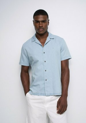 Hombre con camisa azul claro de manga corta y pantalones blancos, de pie frente a un fondo blanco liso.