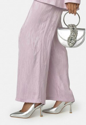 Persona que lleva pantalones anchos texturizados de color lavanda, tacones plateados con tira trasera, sosteniendo un bolso plateado con asa circular y diseño de pedrería.