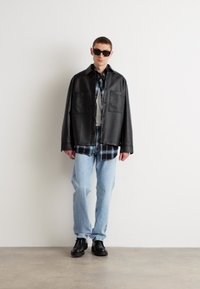 Camisa overshirt de cuero negro con dos bolsillos en el pecho, superpuesta sobre una camisa de cuadro, combinada con jeans azul claro y zapatos negros. Gafas de sol negras puestas.