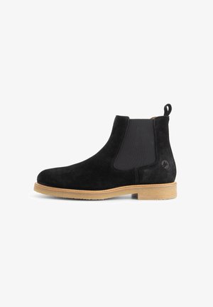 Zwarte suède Chelsea-boot met een tan rubberen zool, elastische zijpaneel en een trekhoekje aan de achterkant, weergegeven in profiel op een witte achtergrond.