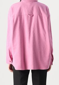 Pink, oversized flannel shirt with a collar, back loop detail, and buttoned cuffs, featuring a soft, textured fabric and dropped shoulders.