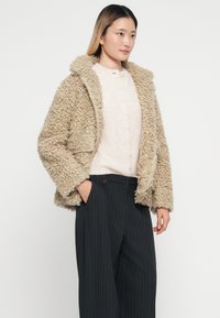 Giacca beige fluffy con texture e cappuccio, tasche frontali, abbinata a un maglione chiaro e pantaloni a strisce nere.