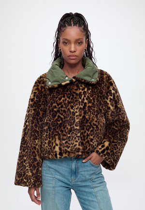 Mulher com cabelo trançado usando um casaco de pelo falso com estampa de leopardo, gola verde e jeans azuis, em pé contra um fundo simples.