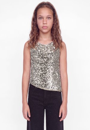 Une fille se tient face à l’objectif devant un fond blanc, vêtue d’un haut sans manches argenté à sequins et d’un pantalon noir.