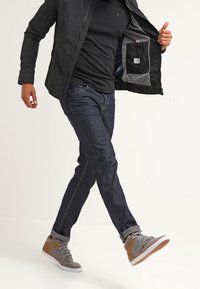 Man i svart jacka, svart skjorta, mörkblå jeans och gråbruna sneakers, som lyfter på jackan för att visa innerfickan.