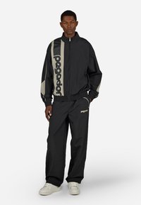 Črna zip-up jakna z svetlo sivo obrobo in velikim logotipom; usklajene črne hlače z logotipom; oboje iz lahkega, teksturiranega materiala.