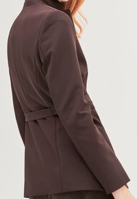 Kobieta w dopasowanej, ciemnobrązowej marynarce z paskiem w talii, pokazana z boku i z tyłu na jednolitym jasnym tle.