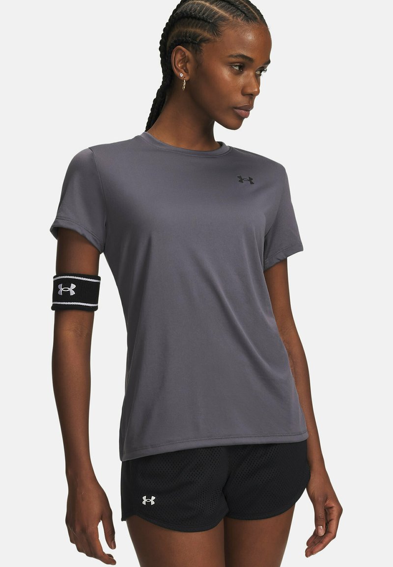 Athletisches Kurzarmshirt aus grauem Stoff mit glatter Textur; verfügt über ein kleines schwarzes Logo. Kombiniert mit schwarzen Mesh-Shorts, akzentuiert durch ein schwarzes Armband.
