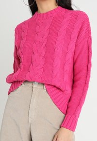 Felle roze gebreide trui met een geribbelde halslijn en kabelpatroon, met zijsplitten en lange mouwen, gecombineerd met beige ribfluweel broek.