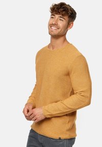 Long-sleeve sweater in mustard yellow, textured knit fabric, crew neckline, fitted sleeves, and subtle stitching details.