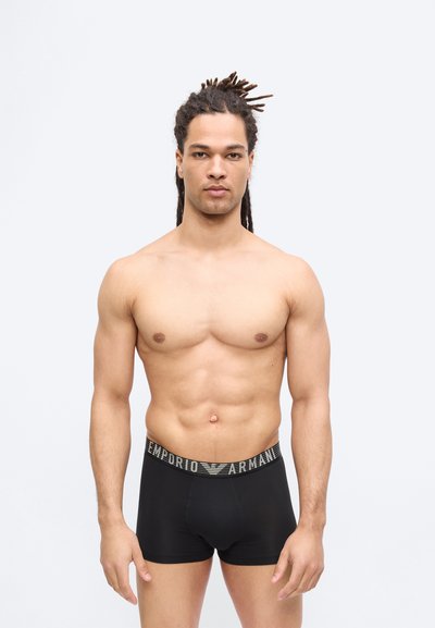 Μυώδης άνδρας με κόμπους στα μαλλιά που στέκεται στραμμένος προς τα εμπρός, φορώντας μαύρα μποξεράκια Emporio Armani, σε απλό φωτεινό φόντο.