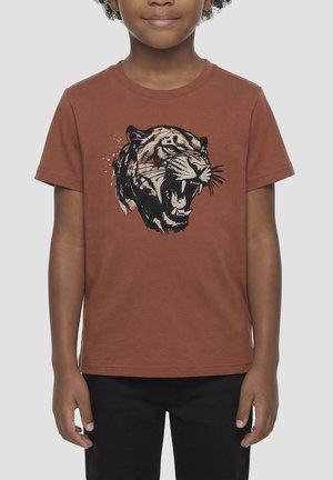 Niño con una camiseta de color óxido con un gráfico detallado de la cabeza de un tigre rugiendo y pantalones negros, de pie contra un fondo liso.