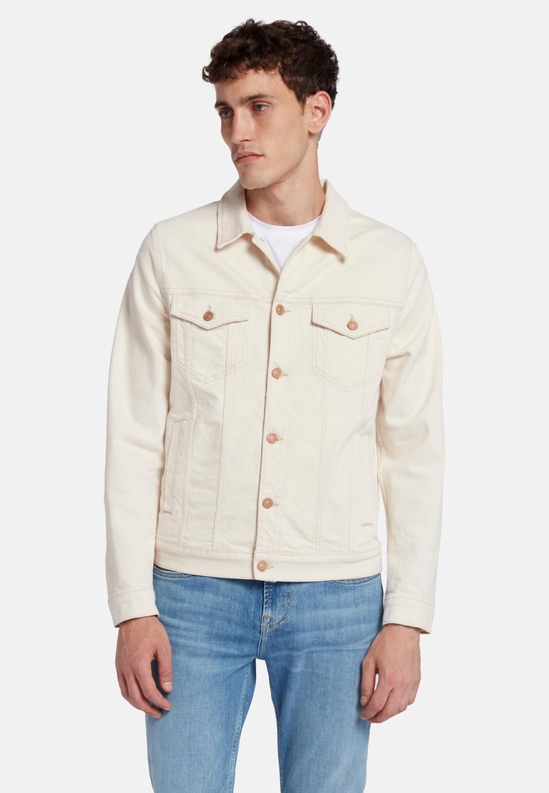 Giacca di jeans chiara con colletto, due tasche sul petto, bottoni in rame e tasche laterali a fessura. Indossata sopra una camicia bianca.